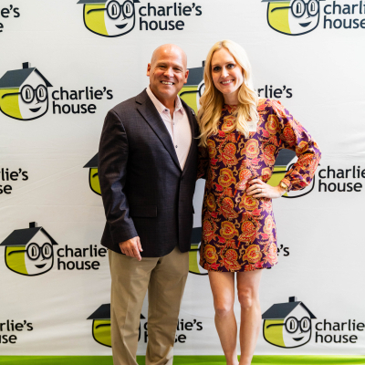 Man in a dark blazer and a woman in a patterned dress posing at the Charlie's House backdrop
