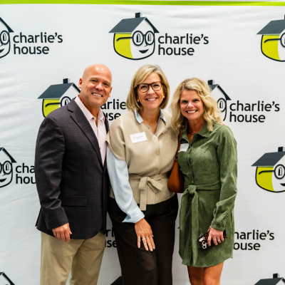 Man in a dark blazer posing with two women at the Charlie's House backdrop
