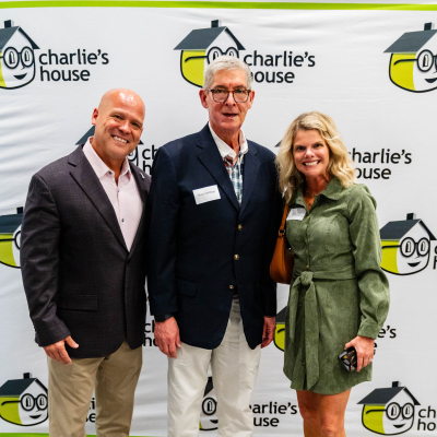 Two men and a woman in green posing at the Charlie's House banner