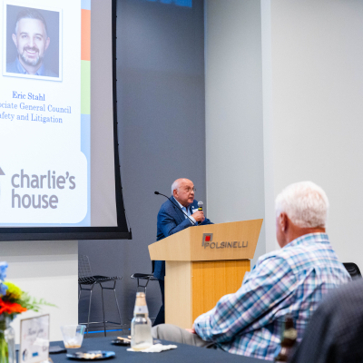 Wide view of the speaker at the podium with panelist photos on the screen behind