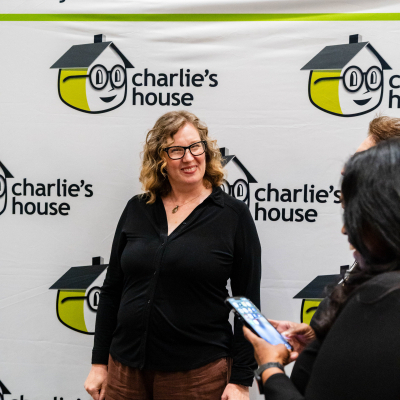 Woman in glasses standing at the Charlie's House backdrop while a guest checks a phone