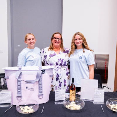 Three women standing behind the raffle table with a cooler bag and bourbon prize