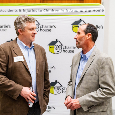 Two men in blazers chatting in front of the Charlie's House banner