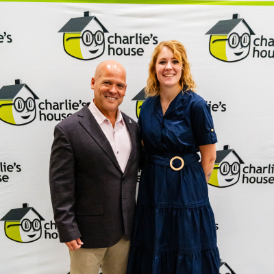 Man in a dark blazer and a woman in a navy dress posing at the Charlie's House banner