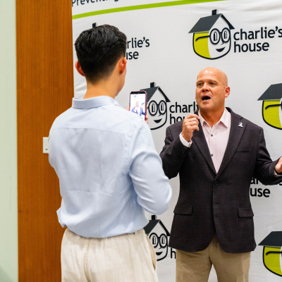 Man gesturing while being recorded on a phone in front of the Charlie's House banner