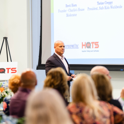 Speaker walking on stage near a HOTS sponsor sign as guests watch
