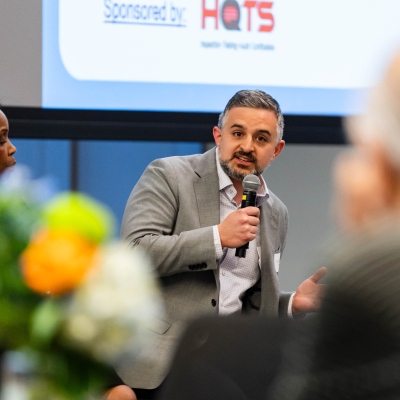 Panelist in a gray blazer speaking into a microphone in front of a HOTS sponsor slide