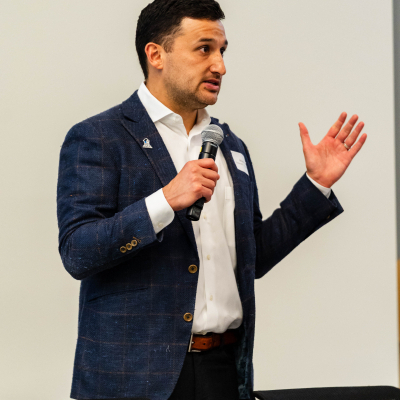 Man in a navy blazer gesturing while speaking into a microphone